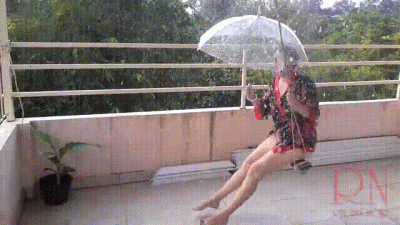 The Lady Is Swinging On A Hanging Swing, Hiding From The Rain Under An Umbrella. Summer Rain In The Garden. Naked Nudist With A Transparent Umbrella On The Terrace